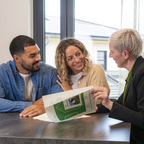 Junges Paar sitzt gemeinsam mit Energieberaterin am Tisch. Die Energieberaterin zeigt dem Paar verschiedenes Infomaterial.