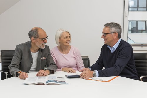 Bild zeigt drei Menschen an einem Tisch mit Informationsmaterialien zur energetischen Sanierung. Rechts im Bild sitzt der Energieberater, links im Bild ein älteres Paar. Sie unterhalten sich.