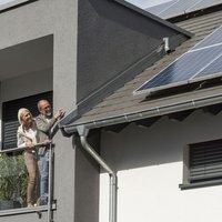 Bild zeigt ein Paar links im Bild, das auf einer Dachterasse steht. Sie schauen auf Photovoltaik-Module auf dem Dach, die sich rechts im Bild befinden.