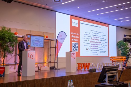 Das Foto zeigt Frank Hettler auf der Bühne des Herbstforum Altbau 2021. Er steht am linken Bildrand, rechts von ihm ist auf der Leinwand eine Wortwolke zu sehen, für die die Teilnehmenden digital abgestimmt haben, worauf sie sich bei der Tagung am meisten freuen. Die Top Begriffe sind "Spannende Vorträge" und "Neue Impulse".