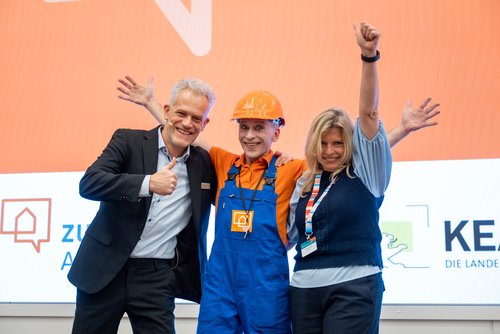 Auf dem Foto sind Frank Hettler (Leiter von Zukunft Altbau), Pablo Zibes (Pantomime und Walking Act) und Natasha Walker (Moderatorin) auf der Bühne beim Herbstforum Altbau 2025 zu sehen. 