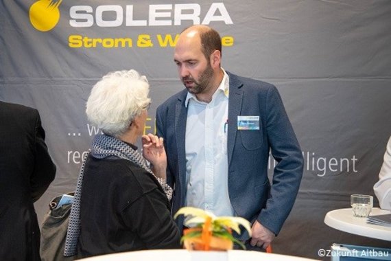 Foto einer Gesprächssituation von zwei Personen vor einer grauen Plakatwand mit gelbem Logo und weißem Solera-Schriftzug. 