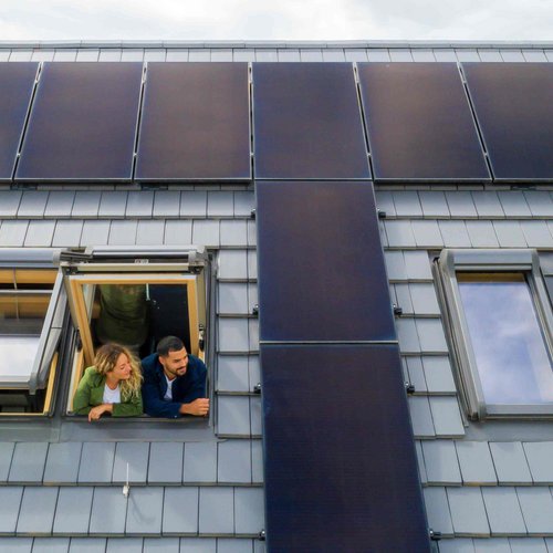 Ein junges Paar steht an einem Dachfenster und schaut hinaus. Um das Fenster herum ist die Dachfläche zu sehen, inklusive Photovoltaikanlage.