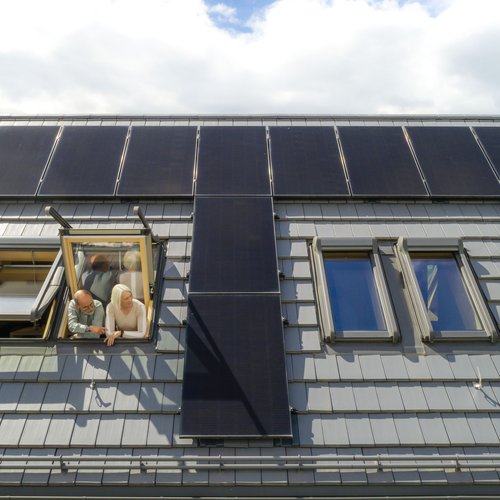 Ein paar schaut zu einem Dachfenster hinaus. Drumherum sind Photovoltaikmodule auf dem Dach zu sehen.