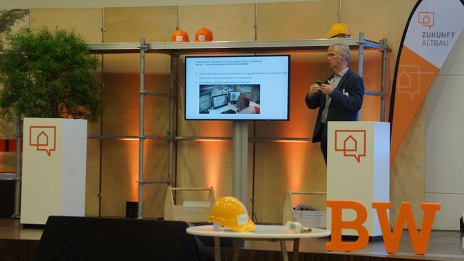 Foto in halbtotaler Einstellung von Dipl-Ing. (FH) Architekt Roland Matzig bei seinem Vortrag auf dem Herbstforum Altbau 2020. Redner rechts im Bild, Präsentation auf Leinwand links daneben.