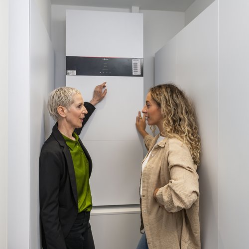 Das Foto zeigt zwei Frauen vor einem Solarstromspeicher. Die Frau links im Bild zeigt auf den Speicher und erklärt der Frau recht im Bild ihre Funktion.