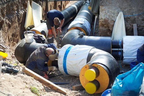 Auf einem Foto sieht man einen offenen Graben mit großen Nahwärmerohren. Zwei Bauarbeiter sind ebenfalls in der Grube und arbeiten gerade an den Rohren.