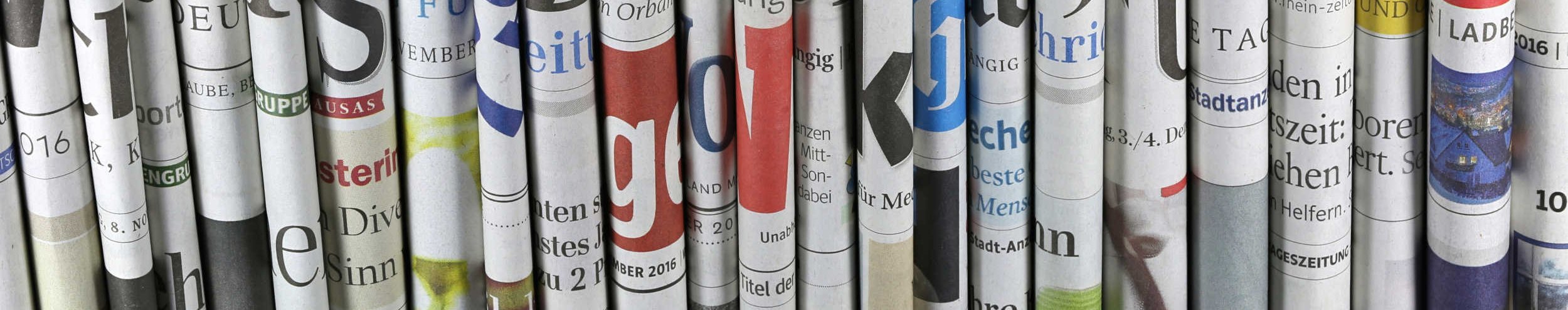 Das Foto zeigt einen Stapel an Magazinen und Zeitungen, das oberste ist aufgeschlagen. Im Hintergrund links ist eine Kaffeetasse erkennbar. 