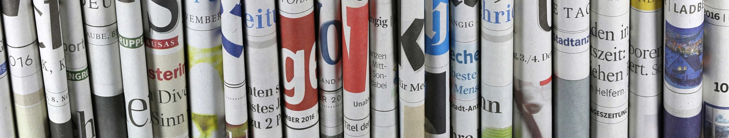 Das Foto zeigt einen Stapel an Magazinen und Zeitungen, das oberste ist aufgeschlagen. Im Hintergrund links ist eine Kaffeetasse erkennbar. 