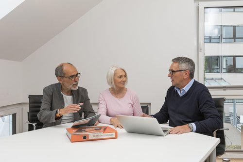 Das Bild zeigt drei Menschen an einem Tisch mit Informationsmaterialien zur energetischen Sanierung. Rechts im Bild sitzt der Energieberater, links im Bild ein älteres Paar. Sie unterhalten sich.