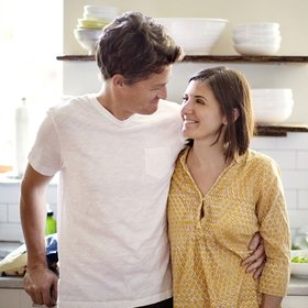 Das Bild zeigt ein Paar in einer Küche in einer halbnahen Einstellung. Sie stehen der Kamera zugewandt und blicken einander an. Der Mann in weißem T-Shirt steht links und hält eine in gelb gekleidete Frau rechts neben sich im Arm.