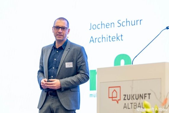 Foto von Architekt Jochen Schurr auf der Bühne links im Bild, rechts neben ihm befindet sich ein weißes Rednerpult mit Zukunft Altbau. 