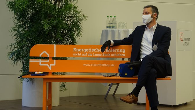 Foto in halbtotaler Einstellung von M.Sc Tobias Nusser von EGS-plan GmbH, sitzend auf einer orangenen Bank auf der Bühne des Herbstforums Altbau 2020. 