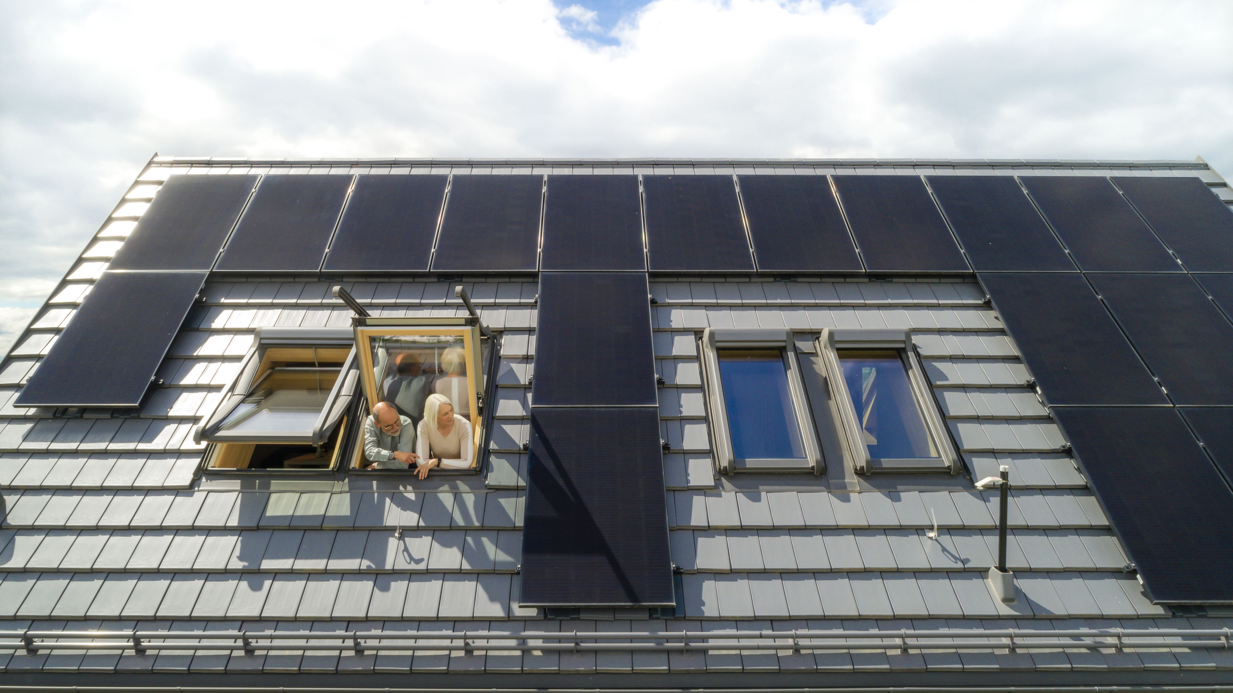 Ein paar schaut zu einem Dachfenster hinaus. Drumherum sind Photovoltaikmodule auf dem Dach zu sehen.