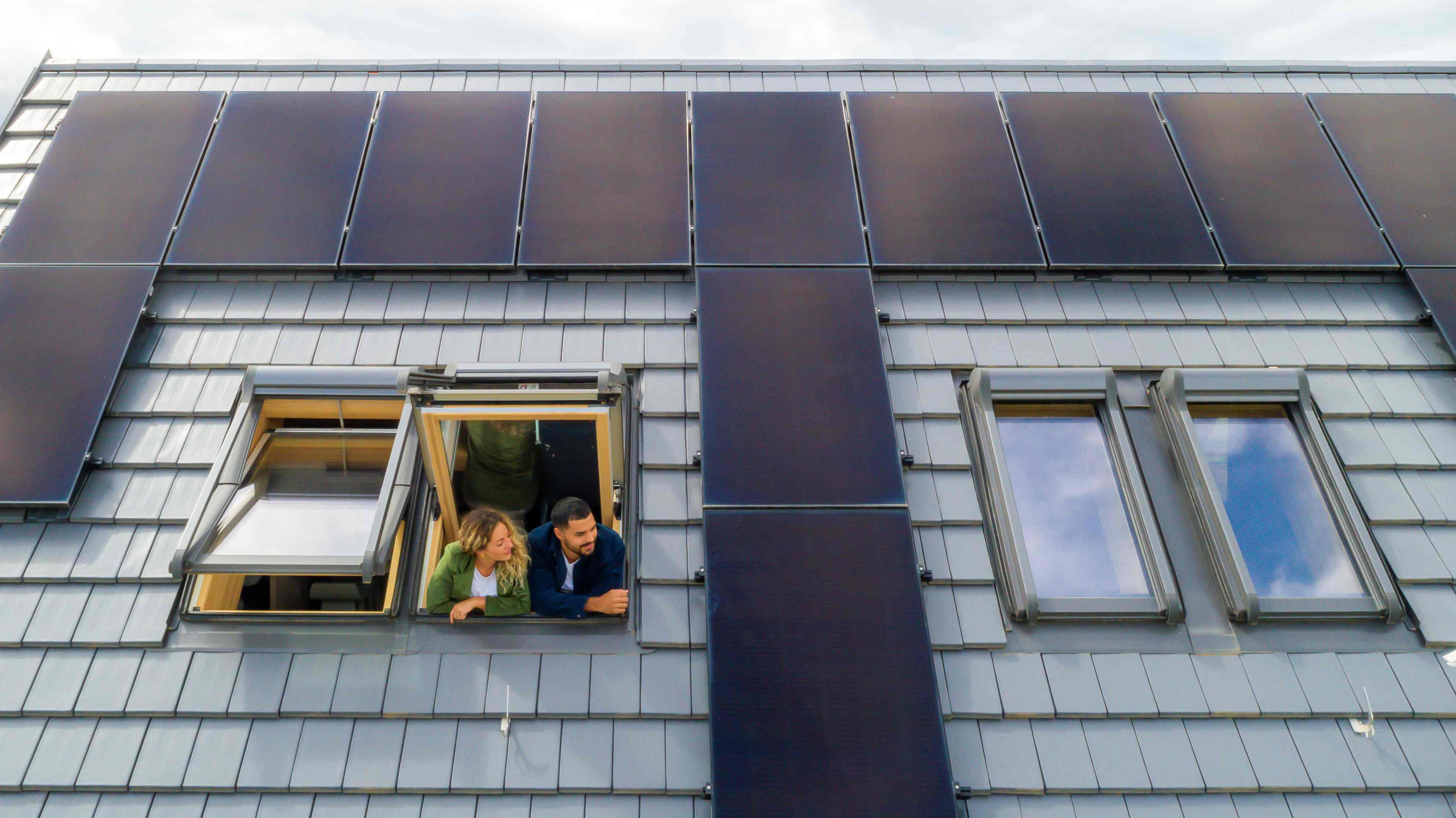 Ein junges Paar steht an einem Dachfenster und schaut hinaus. Um das Fenster herum ist die Dachfläche zu sehen, inklusive Photovoltaikanlage.