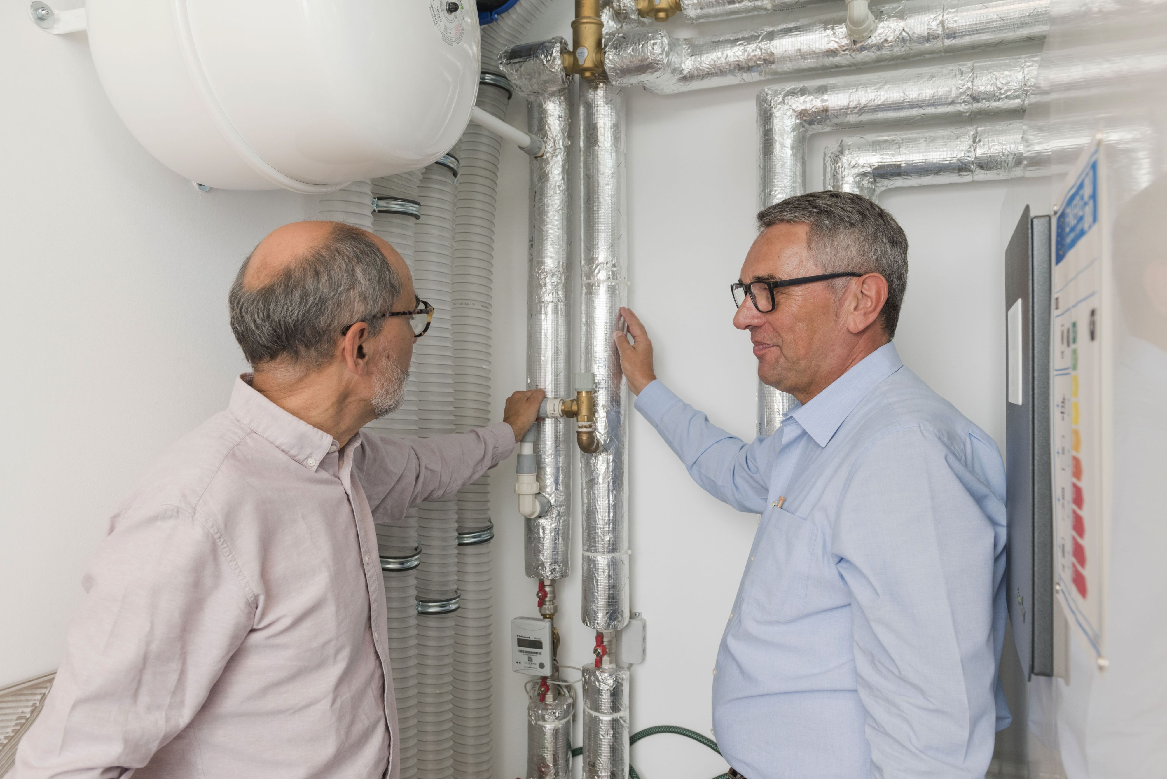 Zwei Männer stehen im Heizungsraum eines Gebäudes. Der Energieberater zeigt dem Eigentümer die Funktionsweise der Heizung.