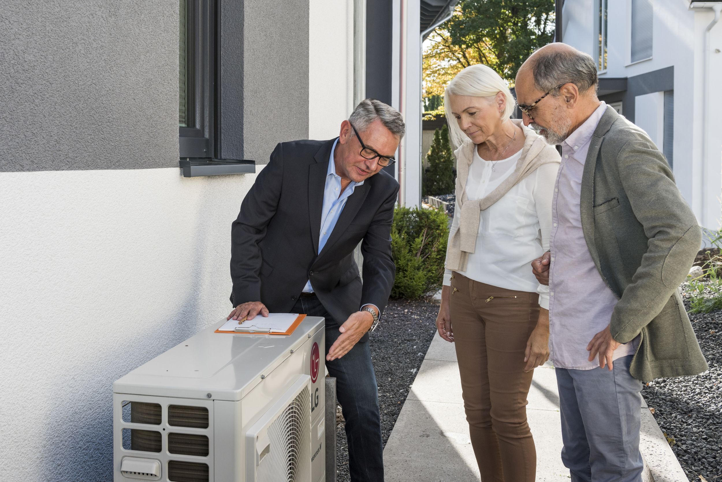 Bild zeigt drei Personen, die vor einer Luft-Wärmepumpe stehen. Der Energieberater links im Bild zeigt darauf und erklärt einem älteren Paar rechts im Bild, wie das Gerät funktioniert.