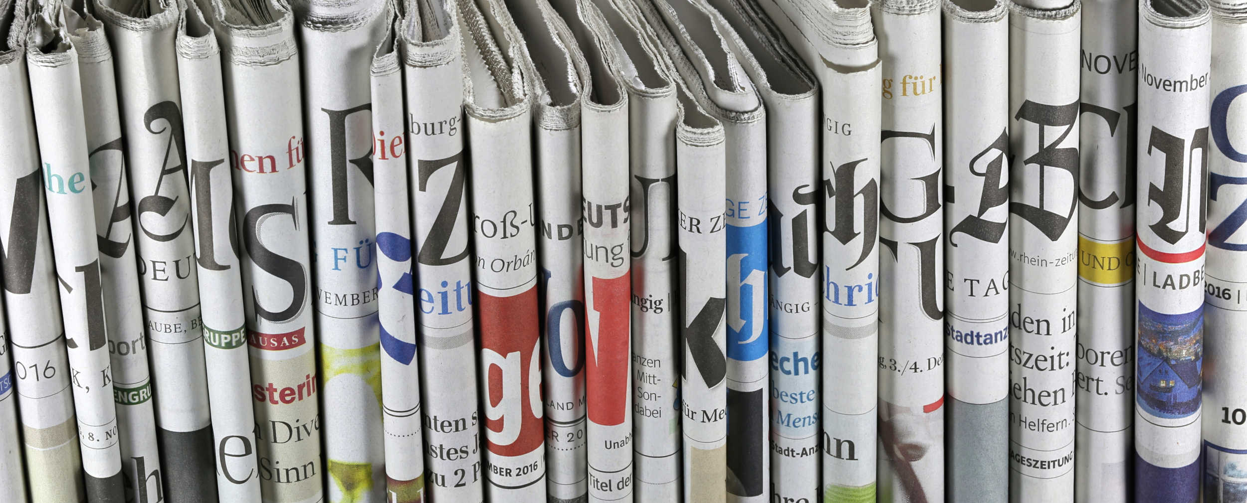 Das Foto zeigt einen Stapel an Magazinen und Zeitungen, das oberste ist aufgeschlagen. Im Hintergrund links ist eine Kaffeetasse erkennbar. 