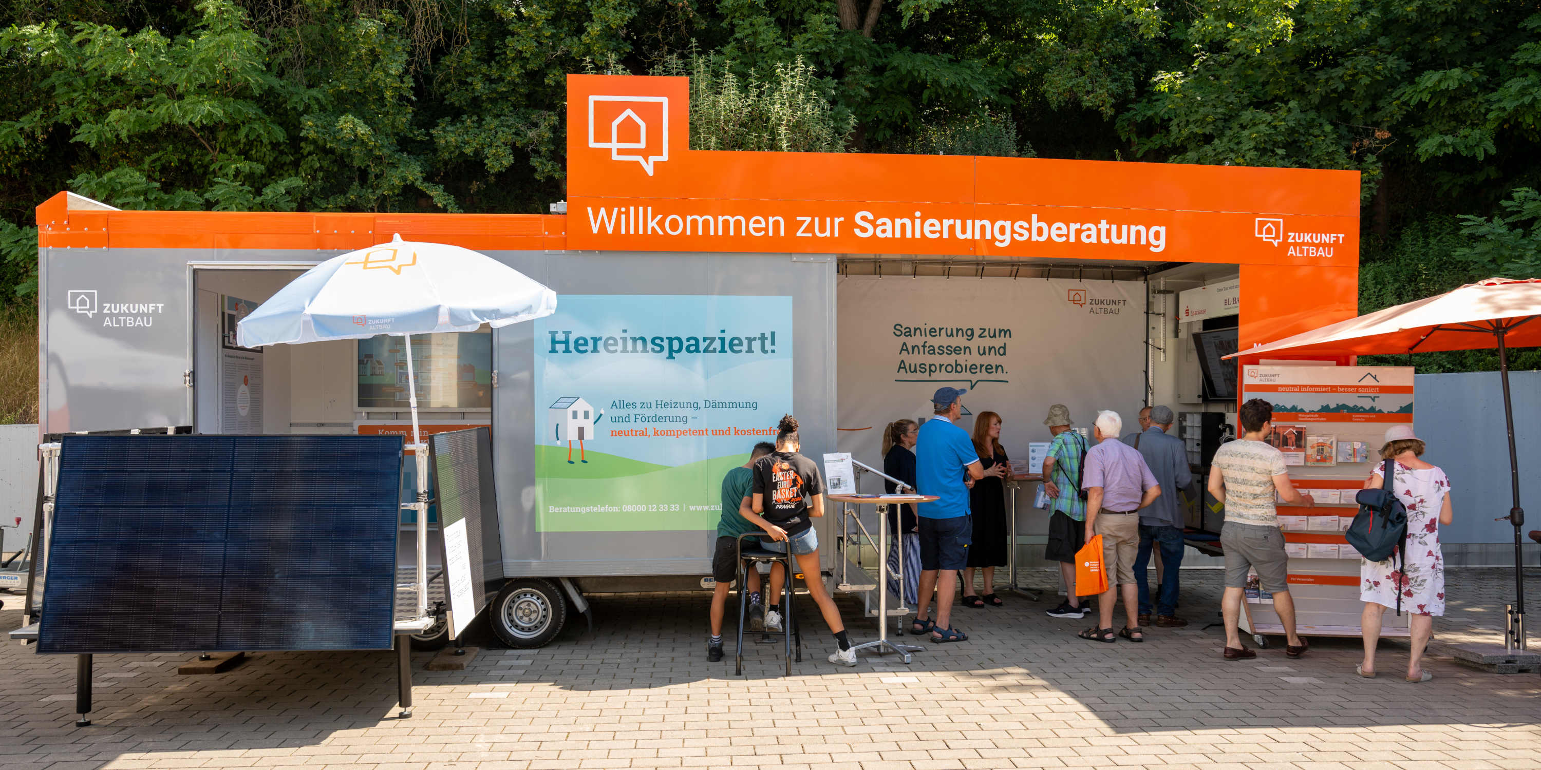 Informationsstand des Sanierungsmobils von Zukunft Altbau im Freien. Der aufklappbare Anhänger in Orange und Grau trägt die Aufschrift „Willkommen zur Sanierungsberatung“. Mehrere Personen informieren sich an Informationsständen und Prospektständern. Im Vordergrund sind Solar- bzw. Photovoltaikmodule aufgebaut.