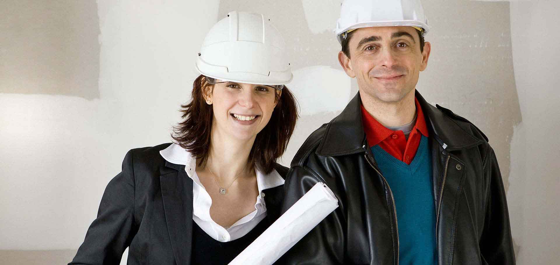 Das Foto zeigt eine Frau und einen Mann mit weißen Bauhelmen in einem halbnahen Bildausschnitt. Die links stehende Frau hält einen zusammengerollten Bauplan in der Hand.